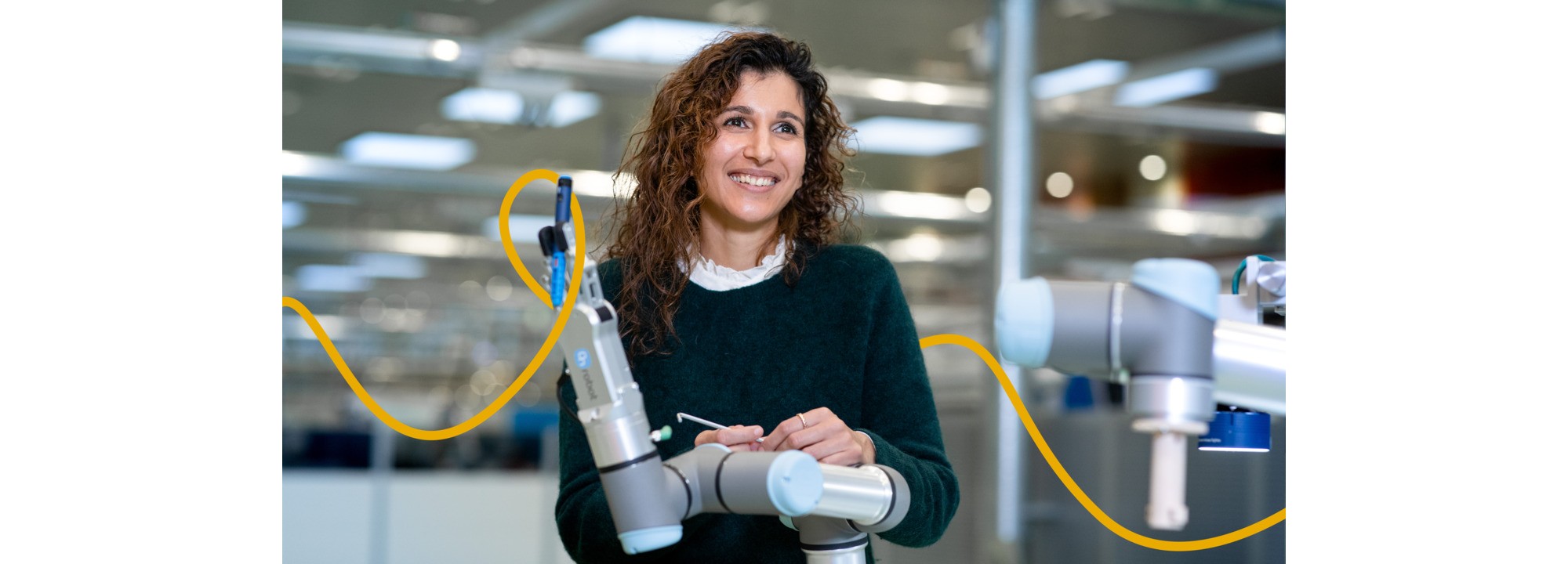 Roa is standing in front of a robot arm. She is holding a tool in her hand, used to adjust the robot arm.