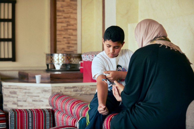 Meshari is in his house with his mother. She is helping him inject his haemophilia medication.