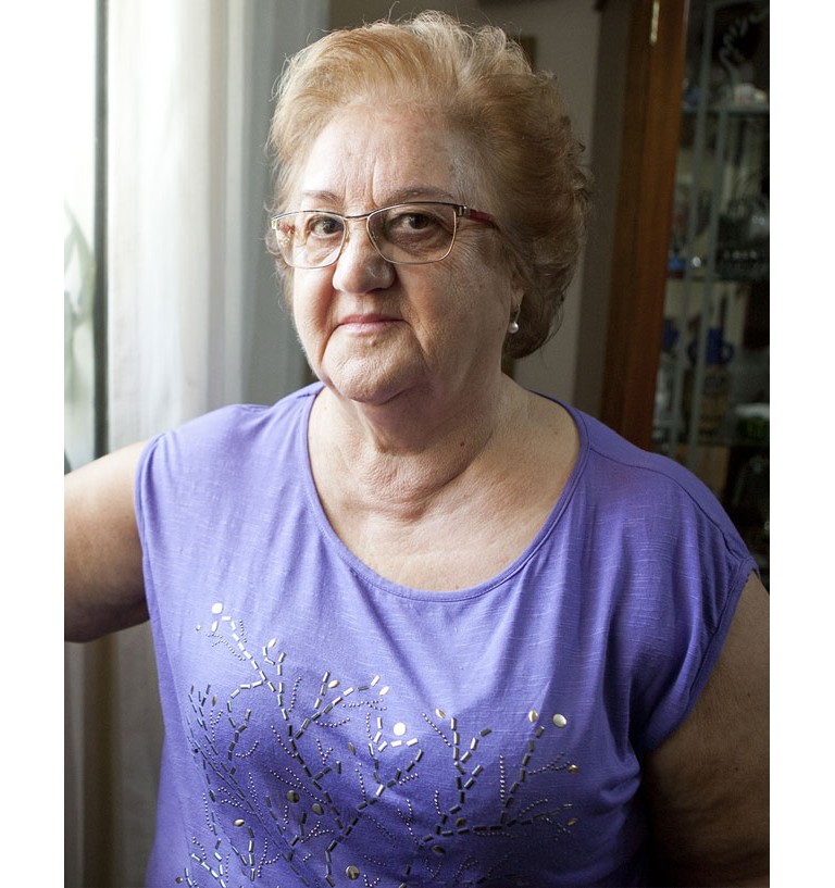 Maria is in her home in Brazil. She is wearing a purple blouse and looks directly into the camera.