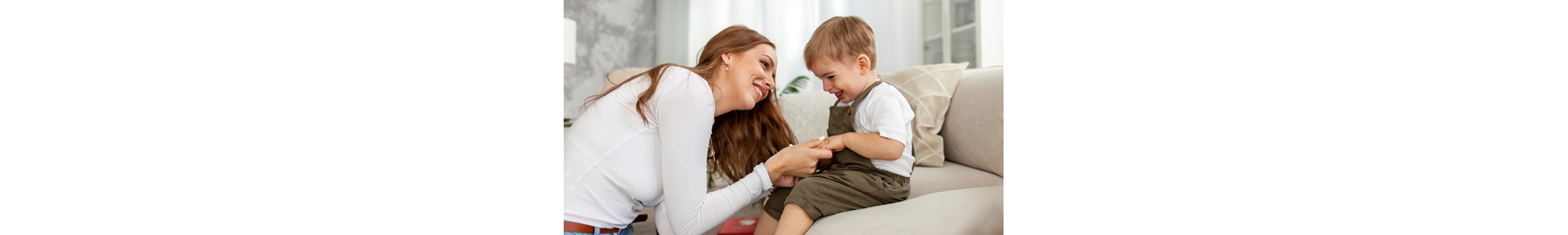 young mother holds the hands of her little son and smiles, 2-year-old boy sits on the sofa with his parent, woman raises and takes care of the child